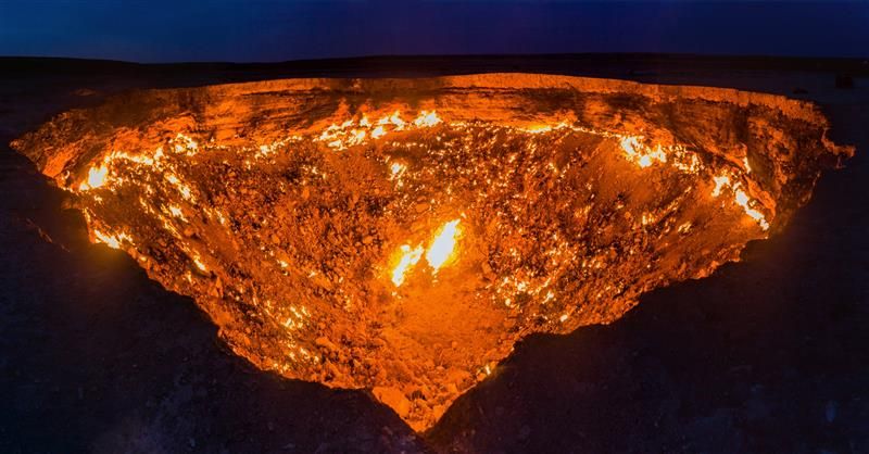 A photo taken at night of the Darvaza crater, showing a large inverted triangle space that is glowing with fire. There are patches of the cater that are brighter than others, while the upper rim of the hole has a near unbroken line of fire going around it.