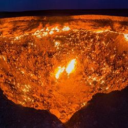 A photo taken at night of the Darvaza crater, showing a large inverted triangle space that is glowing with fire. There are patches of the cater that are brighter than others, while the upper rim of the hole has a near unbroken line of fire going around it.