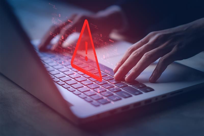 A close up photo of a laptop and two hands manipulating the keys. A large red warning symbol has been superimposed over the keyboard to suggest dangers. 