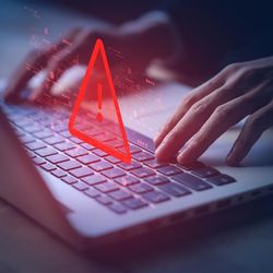 A close up photo of a laptop and two hands manipulating the keys. A large red warning symbol has been superimposed over the keyboard to suggest dangers. 