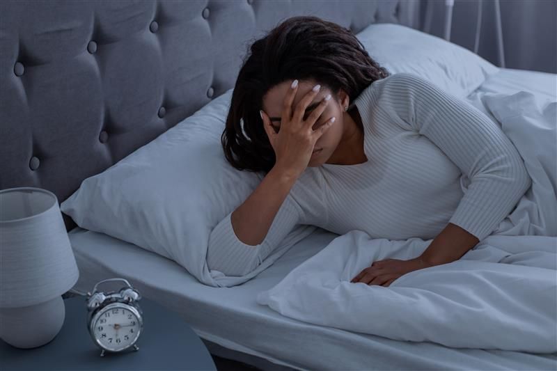 A photo of a woman wearing a white night shirt cradling her face as she lays in bed. The clock on the side of the bed stand shows the time to be 6:15