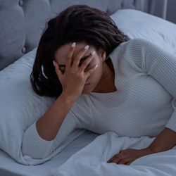 A photo of a woman wearing a white night shirt cradling her face as she lays in bed. The clock on the side of the bed stand shows the time to be 6:15