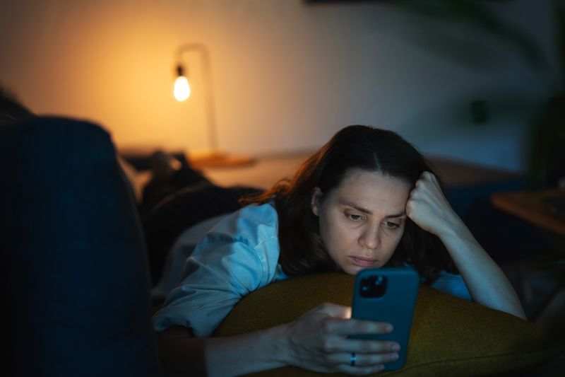 A young woman is lying on her stomach on a sofa. She is staring at her phone and resting her head on her hand. She has a worried look on her face. The room is dark, showing the power of the screen's illumination even though there is a soft yellow light in the background that is coming from a lamp.