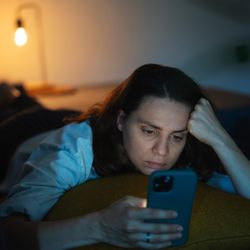 A young woman is lying on her stomach on a sofa. She is staring at her phone and resting her head on her hand. She has a worried look on her face. The room is dark, showing the power of the screen's illumination even though there is a soft yellow light in the background that is coming from a lamp.