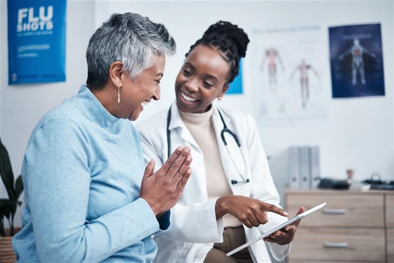 A Black female doctor is explaining information from a clipboard to an elderly lady. They are both laughing and the older lady has her hands pressed together in a similar gesture to a prayer. 