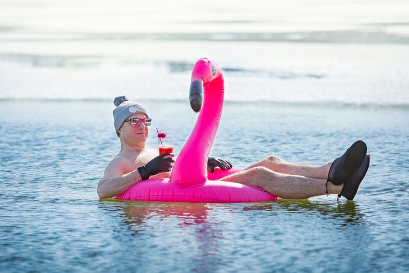 A man swimming in an ice hole in winter in Finland, floating on a pink inflatable flamingo with cocktail in hand. Vacation options, dreaming of summer.