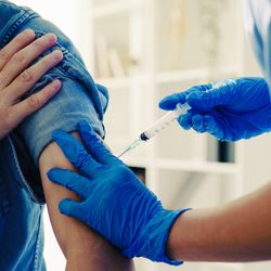 close up of medical professional wearing scrubs and blue gloves administering vaccine into arm of patient wearing blue chambray shirt and white face mask