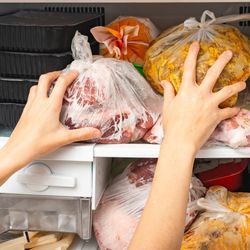 freezer stuffed full of bags of meat and food containers, with person's hands shown pushing more items in