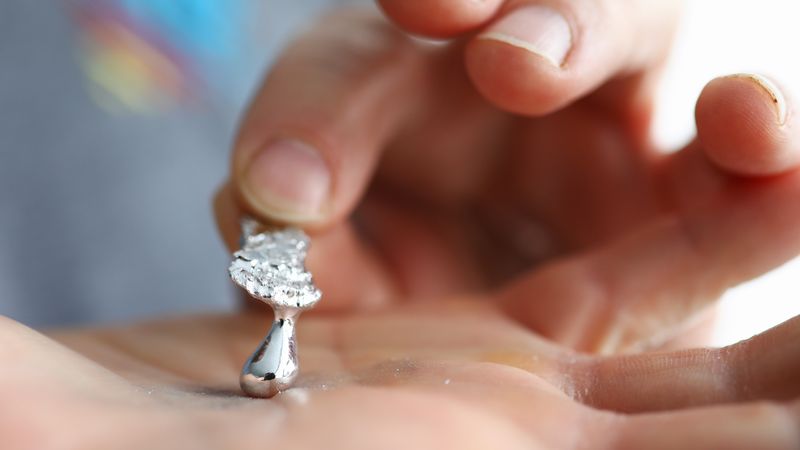 a hand is holding a piece of gallium already turning liquid, a second hand is picking up the solid part showing the liquid drop that formed