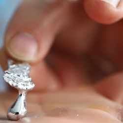 a hand is holding a piece of gallium already turning liquid, a second hand is picking up the solid part showing the liquid drop that formed