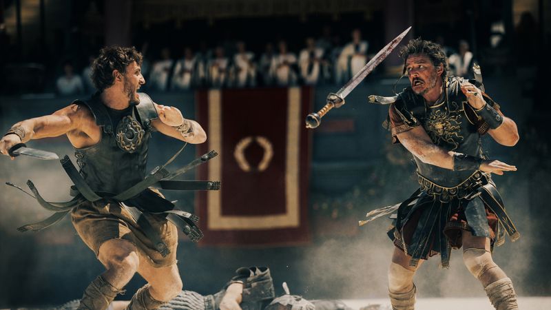 Paul Mescal and Pedro Pascal appear to be duelling in the Roman Colosseum, a still from the motion picture trailer. The two men are wearing leather armour and a sword is hanging in the air, presumably from someone having been disarmed.   