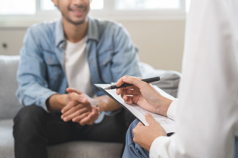 The photo shows a young man sat on a couch talking to a female doctor. The image is cropped so that the top of the man's head and his lower legs are out of frame. He is also blurry. In the right of the foreground, the arms and a folded leg of a female doctor can be seen holding a clipboard and a pen.