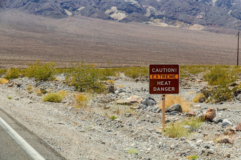 A roadside photo of a "Caution. Extreme Heat. Danger" sign situated in an arid landscape. The sign itself is red with the word "extreme" highlighted by an orange band. In the background, a rocky mountain base can be seen extending beyond the frame.  