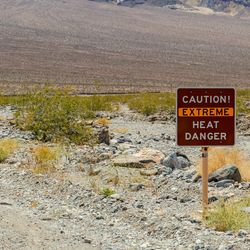 A roadside photo of a "Caution. Extreme Heat. Danger" sign situated in an arid landscape. The sign itself is red with the word "extreme" highlighted by an orange band. In the background, a rocky mountain base can be seen extending beyond the frame.  