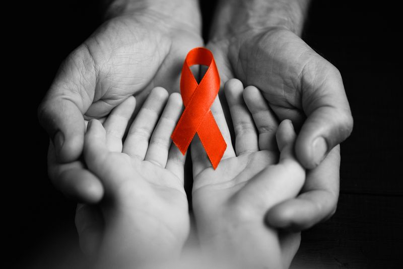 HIV awareness red ribbon held in child's hands, which are held in adult hands. All black and white except the ribbon.