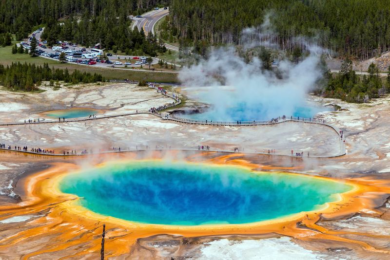 An aerial photo of Yellowstone's Grand Prismatic Spring showing the rich blue of the spring itself as well as the bright yellow and orange stained soils surrounding it. There is steam coming from the water. In the background, another spring is visible which is almost completely covered in thicker plumes of steam. The boardwalks are also visible snaking their way around the springs as tourists visit.