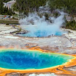 An aerial photo of Yellowstone's Grand Prismatic Spring showing the rich blue of the spring itself as well as the bright yellow and orange stained soils surrounding it. There is steam coming from the water. In the background, another spring is visible which is almost completely covered in thicker plumes of steam. The boardwalks are also visible snaking their way around the springs as tourists visit.