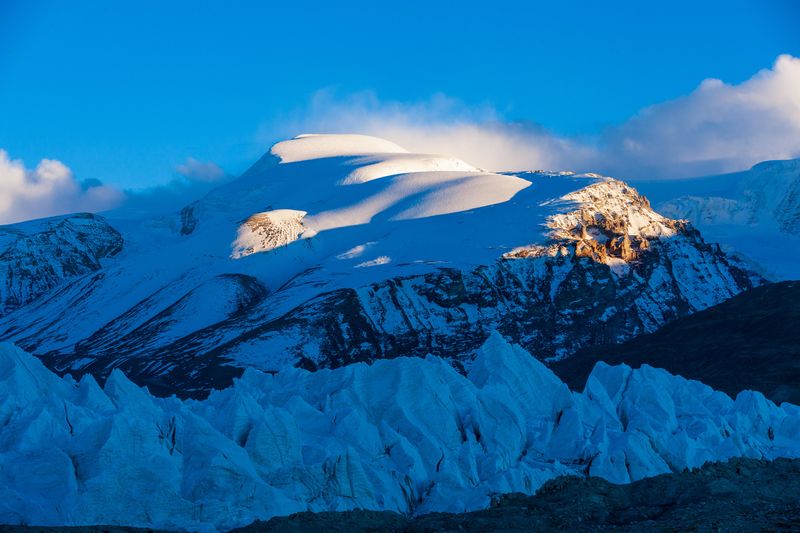 The photo shows the mountain tops and ice around the Tibetan Plateau. In the foreground are jagged ice formations behind which a rock surface is visible which is then covered by snow. Most of the shot is taken in the shade, but there are bumps in the distant snow that are illuminated by soft morning light. 