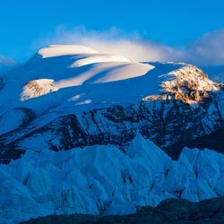 The photo shows the mountain tops and ice around the Tibetan Plateau. In the foreground are jagged ice formations behind which a rock surface is visible which is then covered by snow. Most of the shot is taken in the shade, but there are bumps in the distant snow that are illuminated by soft morning light. 