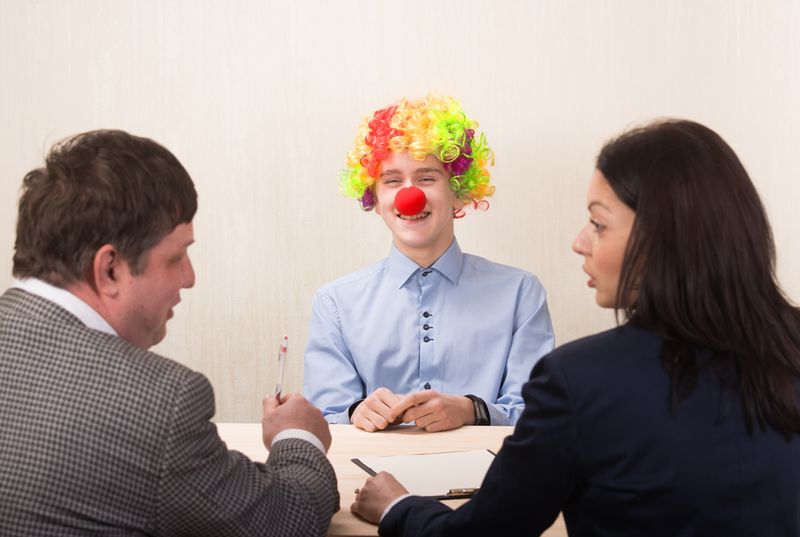 man in clown wig and red nose smiles as male and female interviewers in suits turn to face each other to discuss him