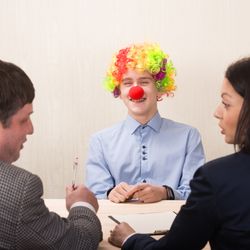 man in clown wig and red nose smiles as male and female interviewers in suits turn to face each other to discuss him