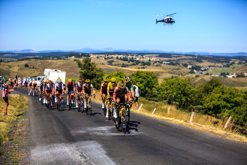 Large group of cyclists during the Tour de France on a road lines with greenery, a helicopter in the blue sky behind them