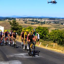 Large group of cyclists during the Tour de France on a road lines with greenery, a helicopter in the blue sky behind them