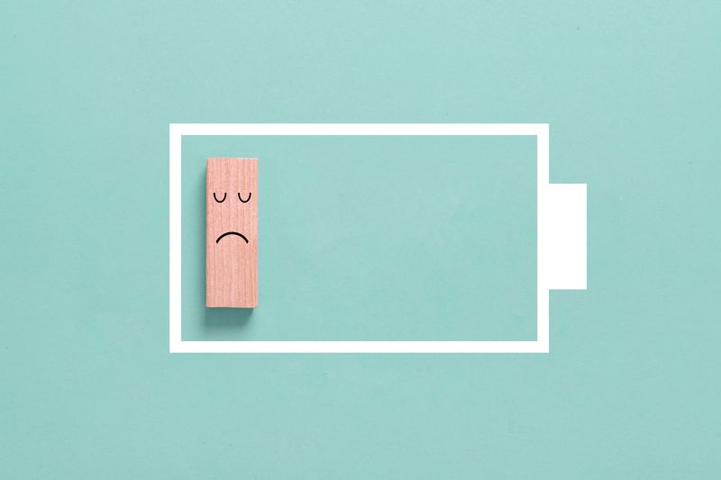 outline of battery in white on a mint green background; a rectangular wooden block with a black sad face is positioned at the end of the battery, suggesting low power