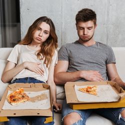 Man and woman sitting on white sofa with pizza leftovers, looking very full.