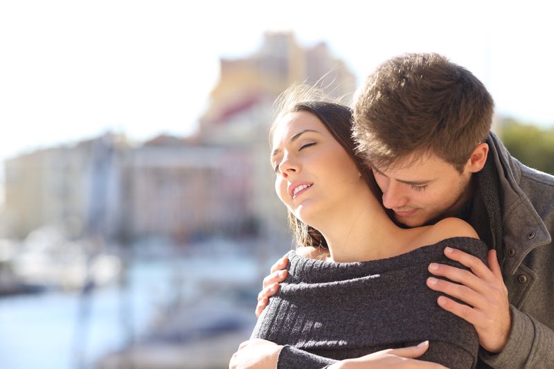 Man standing behind a woman who has her eyes closed, he is holding her shoulders and sniffing her neck