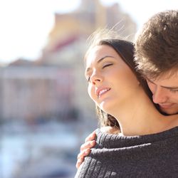 Man standing behind a woman who has her eyes closed, he is holding her shoulders and sniffing her neck