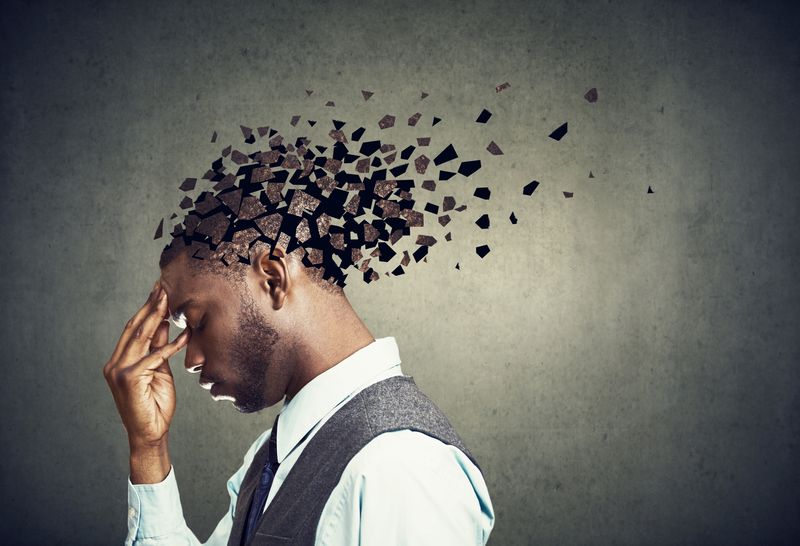 young Black man wearing a suit facing towards the left holding head in hand, with top of head crumbling into pieces to signify memory loss