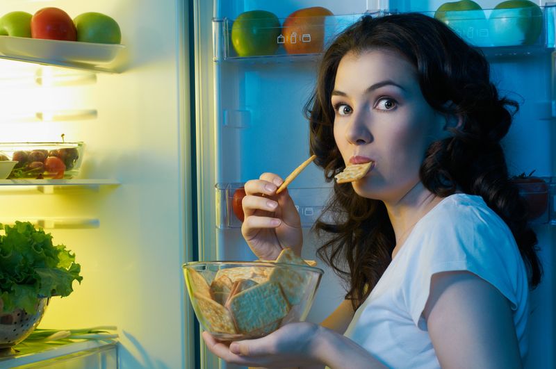 brunette woman looks shocked at camera after being caught eating a bowl of crackers from the fridge; she has a cracker in her mouth