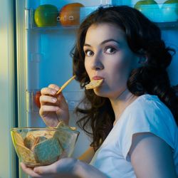 brunette woman looks shocked at camera after being caught eating a bowl of crackers from the fridge; she has a cracker in her mouth