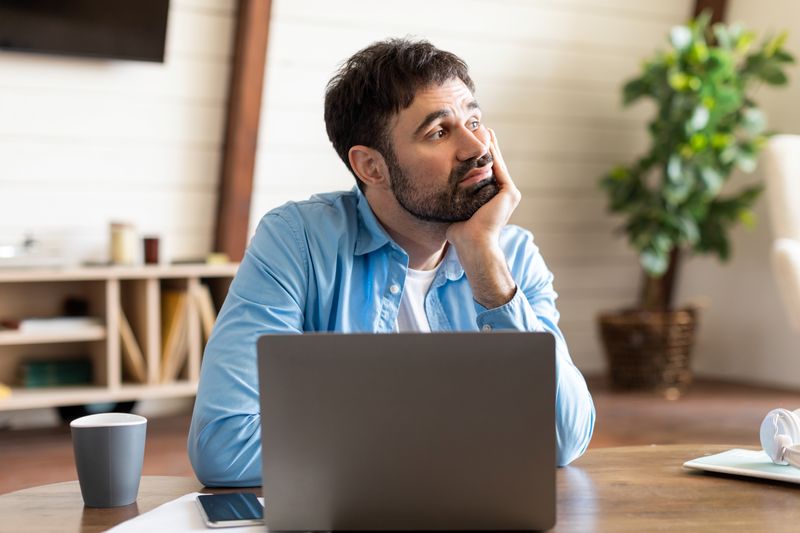 A man is seen deep in thought while sitting at a desk, looking away from his laptop in a well-lit room. His expression reflects contemplation or daydreaming