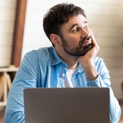 A man is seen deep in thought while sitting at a desk, looking away from his laptop in a well-lit room. His expression reflects contemplation or daydreaming