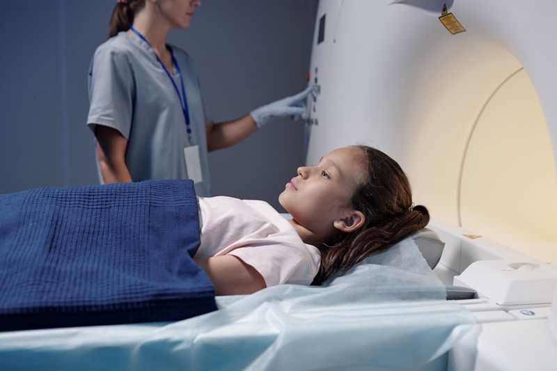 child lying on MRI scanner bed while doctor wearing scrubs in background pushes buttons on the machine