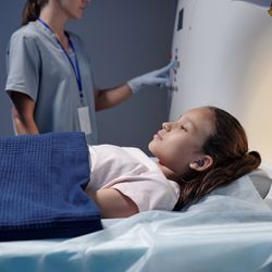 child lying on MRI scanner bed while doctor wearing scrubs in background pushes buttons on the machine