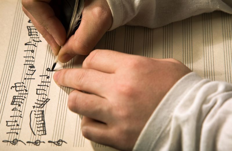 close up of hands as someone writes out music on a sheet of manuscript paper using a quill pen