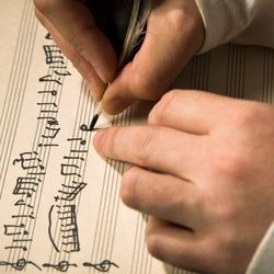 close up of hands as someone writes out music on a sheet of manuscript paper using a quill pen