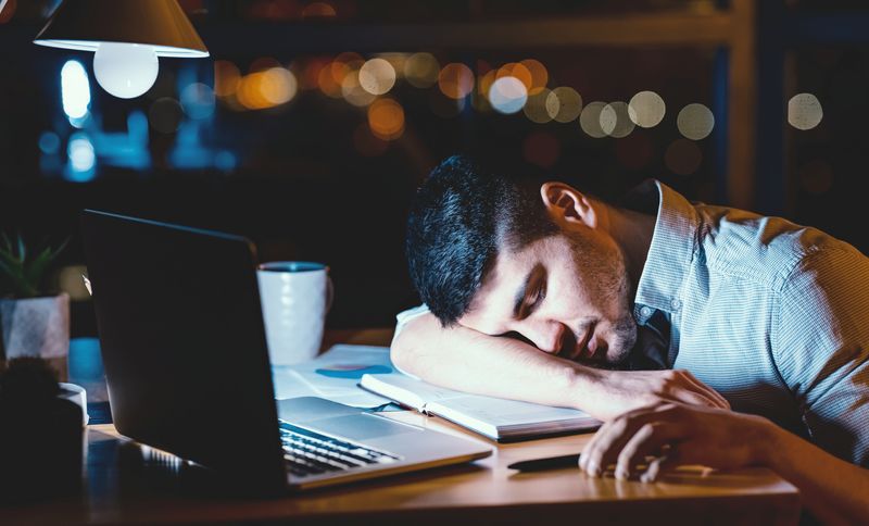 man napping with his head on the desk at work, with laptop, mug and lamp; the sky outside is dark