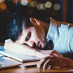 man napping with his head on the desk at work, with laptop, mug and lamp; the sky outside is dark