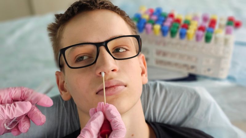 young man with black rectangular glasses lying down while someone takes a swab from his nostril wearing pink gloves; there is a tray of medical containers with multicoloured lids in the background