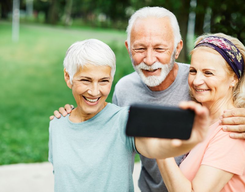 three senior people, two women and one man, smiling together and taking a selfie in a park, wearing exercise clothes