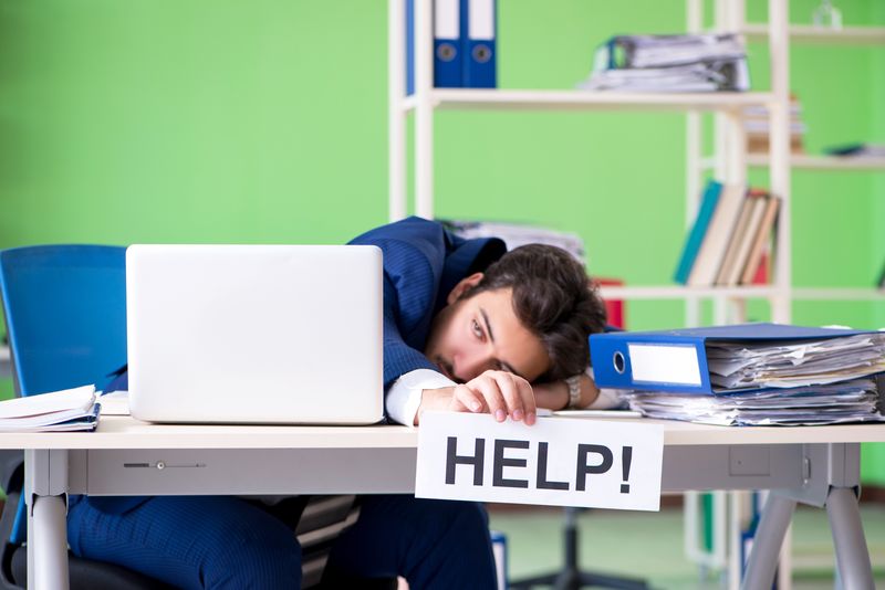 man sitting at desk in office environment with a bright green wall behind him, he is wearing a blue suit and has his head on the desk near a laptop and a pile of folders, and he is holding a sign that says "Help!"