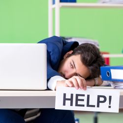 man sitting at desk in office environment with a bright green wall behind him, he is wearing a blue suit and has his head on the desk near a laptop and a pile of folders, and he is holding a sign that says "Help!"