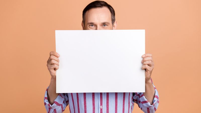 Photo of person holding large piece of blank paper in front of their face, against an orange background