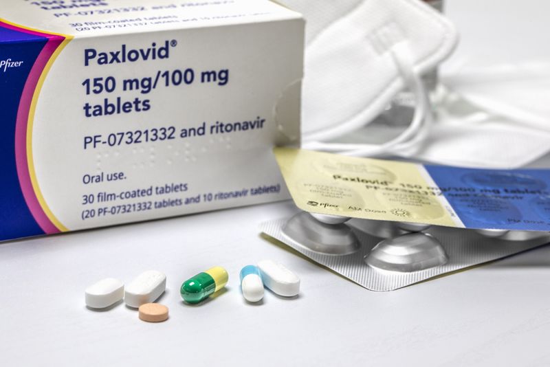 Paxlovid box on a table, with some open blister packs and some N95 masks in the background, and a selection of different coloured pills and capsules in the foreground. 