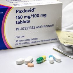 Paxlovid box on a table, with some open blister packs and some N95 masks in the background, and a selection of different coloured pills and capsules in the foreground. 