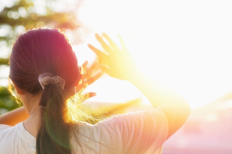 Person with dark hair in a ponytail holding their hands up to shield themselves from the bright sun